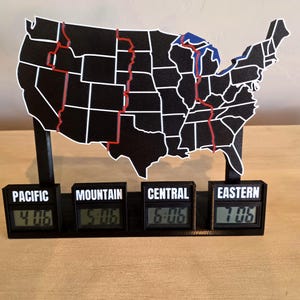 May include: A black map of the United States with white borders and red time zone lines, mounted on a black base with four digital clocks labeled Pacific, Mountain, Central, and Eastern, displaying the times 4:06, 5:06, 6:06, and 7:06 respectively.