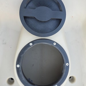 May include: A circular black plastic hatch cover sits above an open, matching circular hatch frame mounted on a beige surface. The frame features six small screw holes around its perimeter and a small directional arrow icon at the bottom edge.
