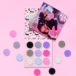 Może przedstawiać: Paleta cieni do powiek w stylu pastel goth z lusterkiem i pasującą ilustracją. Paleta zawiera dziewięć kolorów cieni do powiek i kilka luźnych cieni do powiek w kolorze różowym, fioletowym i czarnym. Ilustracja przedstawia dziewczynę z niebieskimi włosami.