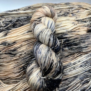 May include: A skein of yarn in a speckled pattern of black, cream, and brown. The yarn is wound into a ball shape and is sitting on a background of the same yarn.