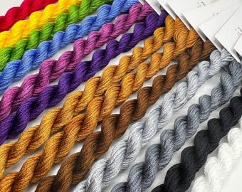 Puede incluir: Una colección de 12 ovillos de hilo de bordar en varios colores, incluyendo rojo, amarillo, verde, azul, morado, dorado, marrón, gris, negro y blanco. El hilo está dispuesto en una fila, con los colores pasando gradualmente de claro a oscuro.