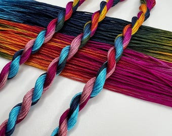 May include: Close-up of colorful embroidery floss in shades of blue, pink, red, orange, and green. Three twisted skeins of floss are in the foreground, with bundles of floss in the background. The floss is used for embroidery and other crafts.