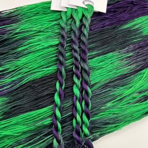 May include: Close-up of embroidery floss in shades of green, purple, and black. Three twisted bundles of floss are in the foreground, with loose floss in the background. The floss is labeled "Pirate + Robin" and "Strand 100% Cotton Embroidery Floss".