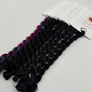 May include: A collection of six-strand embroidery floss skeins arranged in a fan shape. The threads transition from a vibrant magenta and dark grey variegated color to solid black. Each skein is attached to a white paper tag labeled Pirate + Robin.