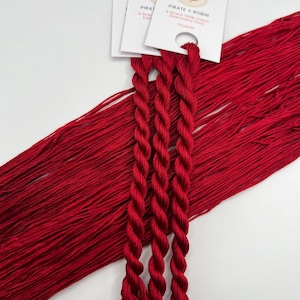May include: Bright red embroidery floss bundles with white labels. The labels read "PIRATE + ROBIN" and "6 Strand 100% Cotton Embroidery Floss, 10 yards". The floss is tightly twisted into three bundles, with loose floss in the background.
