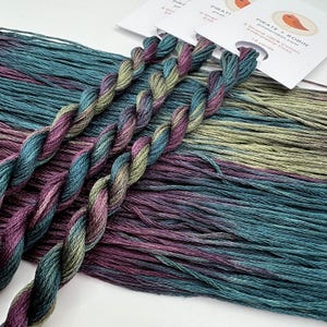 May include: Close-up of embroidery floss skeins in shades of teal, purple, and olive green. The floss is twisted into a rope-like form, with loose strands visible. White labels with a bird logo and text are in the background.