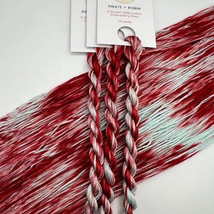 May include: Close-up of embroidery floss in shades of red, white, and light blue. The floss is labeled "PIRATE + ROBIN" and is 10 yards of 6-strand 100% cotton embroidery floss. The floss is arranged in a braided pattern.
