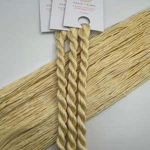 May include: Cream-colored embroidery floss from Pirate + Robin. The image shows three skeins of twisted floss, with the brand's label visible. The label includes the text "6 Strand 100% Cotton Embroidery Floss" and "10 yards". The floss is a light, neutral color.