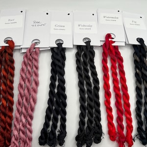May include: A selection of colorful embroidery floss in various shades, including rust, rose, black, red, and gray. Each skein is labeled with a color name and dye code. The floss is arranged in a row, showcasing the range of colors.