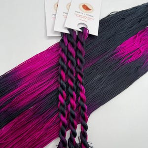 Puede incluir: Tres madejas de hilo de bordar en degradado de negro, rosa y morado, con tres madejas trenzadas a juego en primer plano. La etiqueta indica "Pirate + Robin" con un logotipo de pájaro y 9,14 metros de hilo.