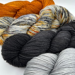 May include: Four skeins of yarn in different colors: a bright orange, a speckled gray and black, a solid black, and a solid gray.