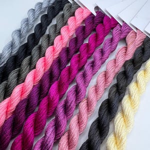 May include: A collection of embroidery floss in various shades of pink, purple, and gray, arranged in a fan shape. The floss is wound on spools and labeled with the brand name "PIR".