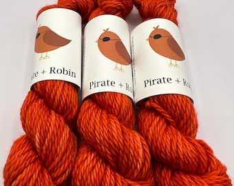 Peut inclure: Trois écheveaux de fil orange avec des reflets blancs. Chaque écheveau est enveloppé d'une étiquette blanche avec un oiseau de dessin animé portant un cache-œil et le texte "Pirate + Robin".