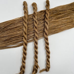 May include: Three braided strands of brown embroidery floss are displayed against a backdrop of loose, similar-colored threads. The floss has a slightly textured appearance, and the color is a warm, earthy brown.
