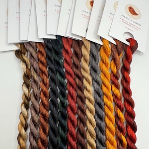 Puede incluir: Un conjunto de 12 carretes de hilo de bordar en varios tonos de marrón, naranja, rojo, verde y gris. Los carretes están etiquetados con "Pirate + Robin" y "6 hebras 100% algodón hilo de bordar 10 yardas".