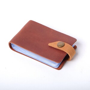 May include: A brown leather card holder with a clear plastic insert and a brown leather strap with a brass button closure.