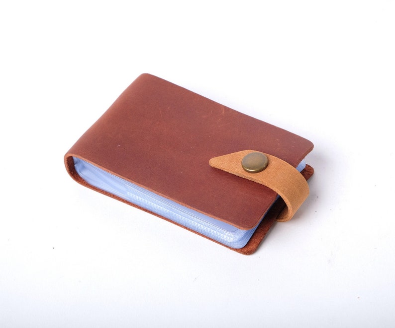 May include: A brown leather card holder with a clear plastic insert and a brown leather strap with a brass button closure.