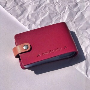 May include: A burgundy leather card holder with a tan leather strap and a silver snap closure. The holder has the text "panikrepto" embossed on the front.