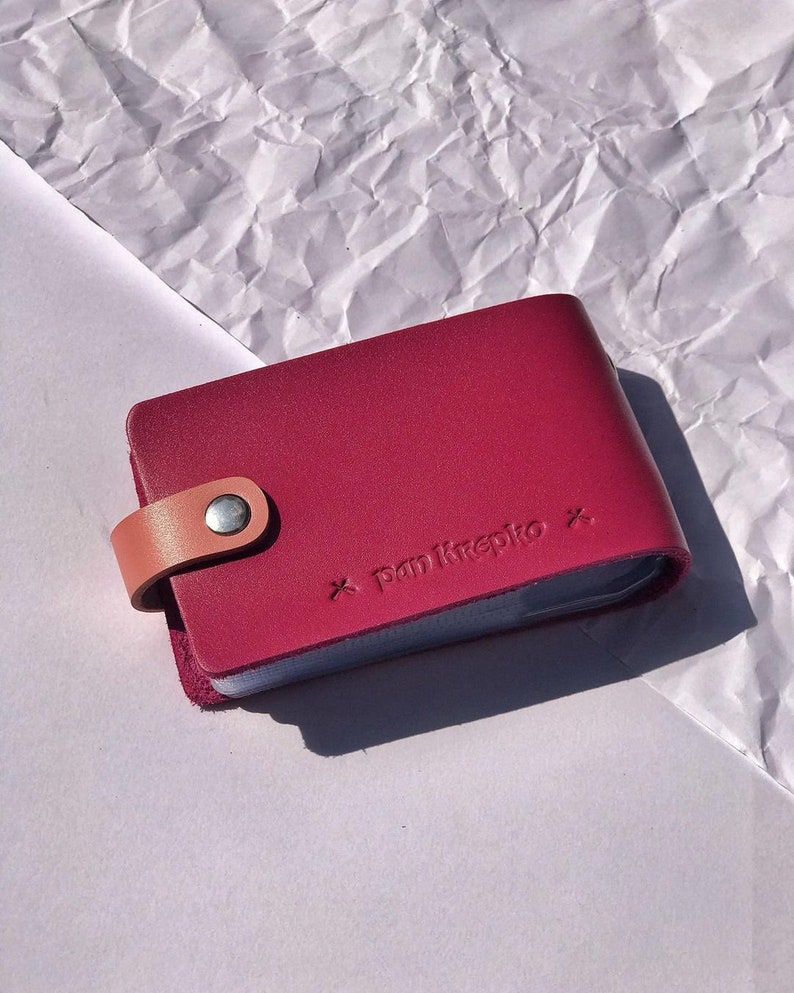 May include: A burgundy leather card holder with a tan leather strap and a silver snap closure. The holder has the text "panikrepto" embossed on the front.