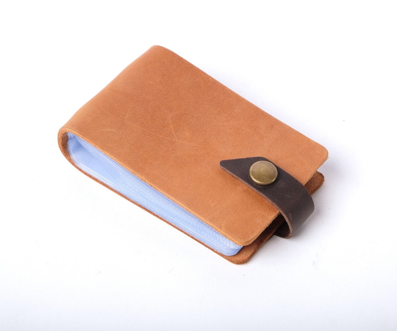May include: A brown leather card holder with a clear plastic insert and a brown leather strap with a brass button closure.