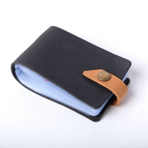 May include: A black leather card holder with a brown leather strap and a brass button closure. The holder has a clear plastic insert for holding cards.