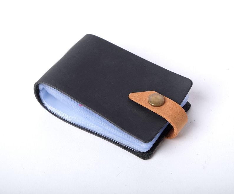 May include: A black leather card holder with a brown leather strap and a brass button closure. The holder has a clear plastic insert for holding cards.