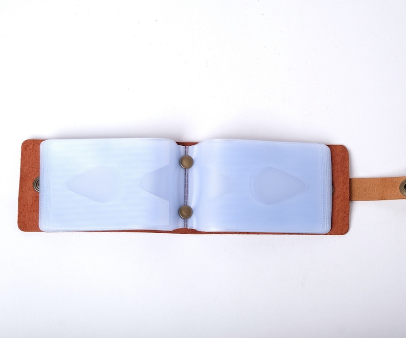 May include: A brown leather card holder with a clear plastic insert for holding cards. The holder has a strap with a metal button.