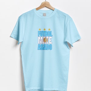 May include: Light blue t-shirt with a graphic print of the words "FUTBOL MATE ASADO" in white and blue letters with a yellow sun and stars. The graphic also includes the blue and white colors of the Argentinian flag.