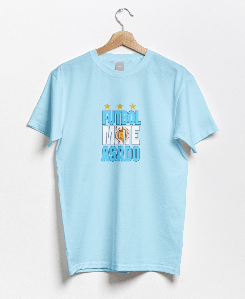 May include: Light blue t-shirt with a graphic print of the words "FUTBOL MATE ASADO" in white and blue letters with a yellow sun and stars. The graphic also includes the blue and white colors of the Argentinian flag.