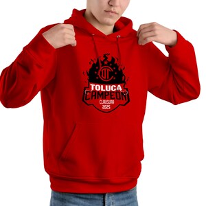 May include: A red hooded sweatshirt with a black graphic that reads "TOLUCA CAMPEON CLAUSURA 2023". The graphic includes a team logo and a flame design. The sweatshirt has a front pocket and a drawstring hood.
