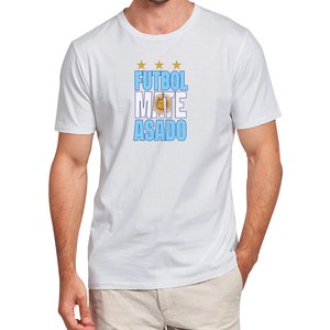 May include: White t-shirt with a blue and white graphic that says "FUTBOL MATE ASADO" with a gold star above the word "FUTBOL" and a gold sun with a circle around it in the middle of the word "MATE".