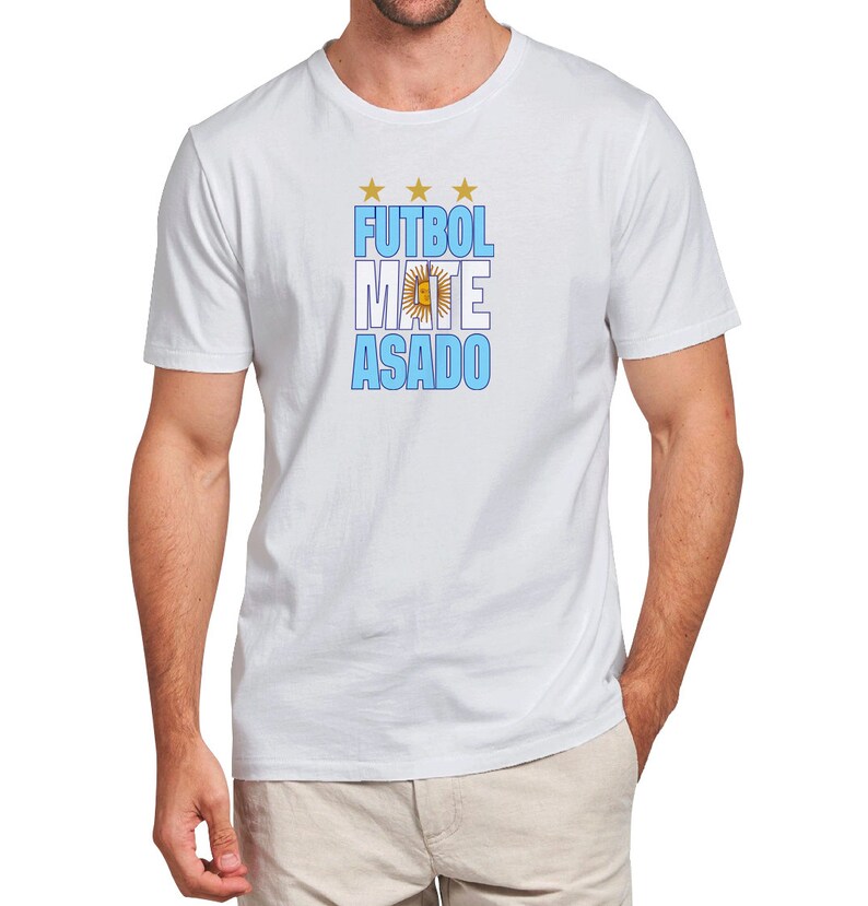 May include: White t-shirt with a blue and white graphic that says "FUTBOL MATE ASADO" with a gold star above the word "FUTBOL" and a gold sun with a circle around it in the middle of the word "MATE".