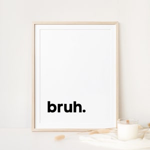 May include: A framed print with the word "bruh" in black text on a white background. The frame is light wood and the print is sitting on a white surface with a white candle and dried grasses.