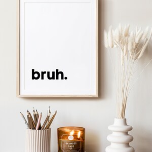 May include: A framed print with the word "bruh" in black text on a white background. A brown candle with a white label that reads "Oakmoss Sage" is sitting on a white book. A vase with a white, textured, wavy design holds dried white pampas grass.