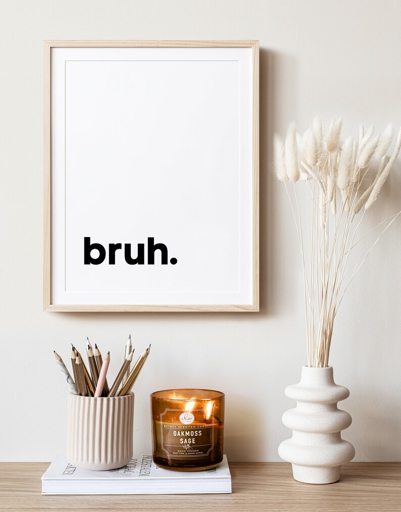 May include: A framed print with the word "bruh" in black text on a white background. A brown candle with a white label that reads "Oakmoss Sage" is sitting on a white book. A vase with a white, textured, wavy design holds dried white pampas grass.