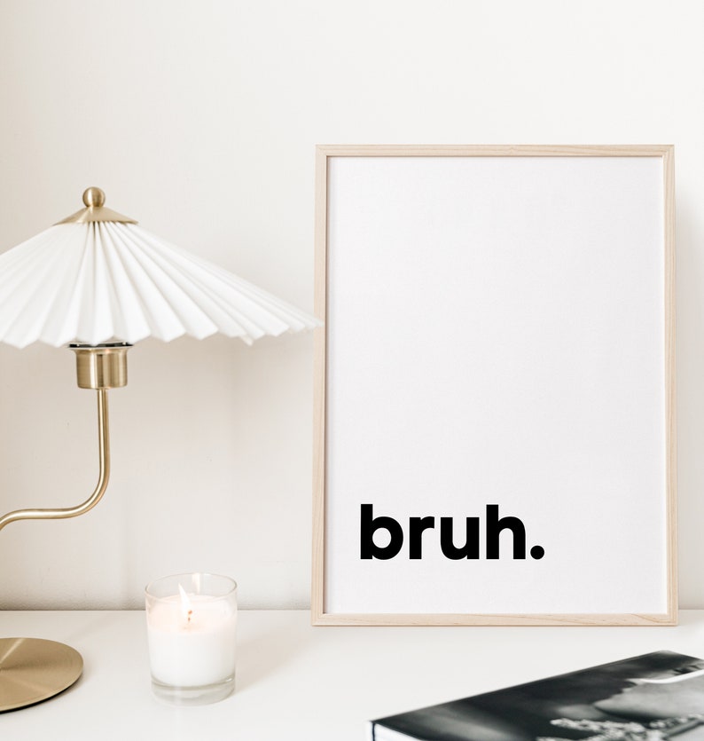 May include: A framed print with the word "bruh" in black text on a white background. The print is in a light wood frame and is sitting on a white surface with a lit candle and a gold lamp.