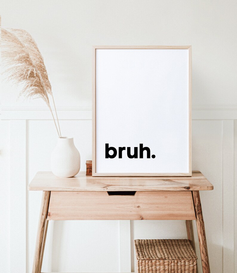 May include: A framed print with the word "bruh" in black text on a white background. The print is sitting on a light wood table with a drawer and a wicker basket underneath.