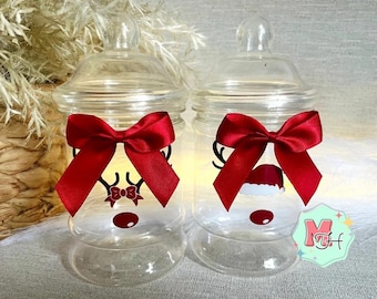 May include: Two clear glass jars with lids, each adorned with a red satin bow. One jar features a reindeer face design with antlers and a bow tie, while the other has a Santa hat design. The jars are festive and decorative.