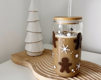 May include: A clear glass tumbler with a bamboo lid and a straw. The tumbler is decorated with gingerbread men and snowflakes. The tumbler is filled with a brown liquid.