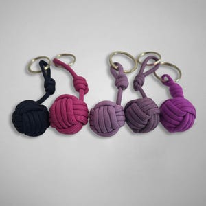 Puede incluir: Cinco llaveros de nudo de puño de mono hechos de cuerda trenzada en tonos negro, rosa oscuro, morado claro, morado medio y morado brillante. Cada llavero presenta un extremo esférico anudado unido a una anilla de metal plateado por un cordón corto.