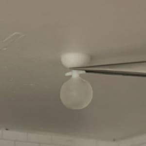 May include: A small white ceiling light fitting with a spherical bulb and a circular base. Tweezers hold the light against a white backdrop. The fitting is likely for a doll's house or model.