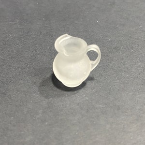 Puede incluir: Una pequeña jarra de vidrio esmerilado con cuerpo redondeado y asa. La jarra es blanca y tiene un acabado liso y mate.
