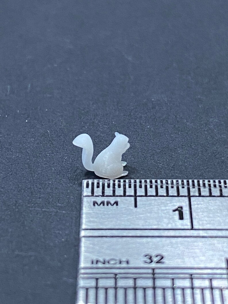 May include: A small, white, 3D printed squirrel figurine, approximately 2.5 cm in length, sitting on a ruler.