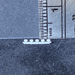 May include: A close-up of a white plastic model of a ship with six small round windows. The model is about 1 inch long and is sitting on a gray surface next to a ruler.