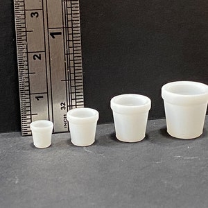 May include: Four white plastic flower pots of varying sizes. The pots are arranged from smallest to largest, with the smallest pot measuring approximately 1 inch in diameter and the largest pot measuring approximately 3 inches in diameter.