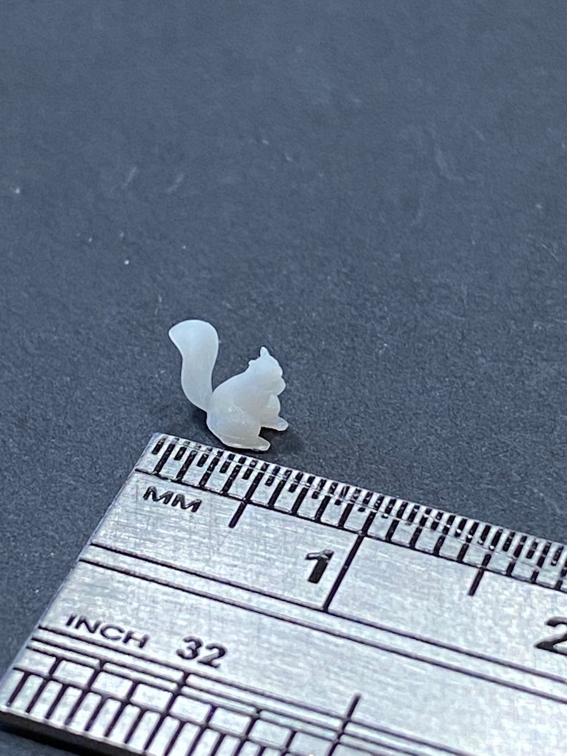 May include: A small, white, 3D printed squirrel figurine measuring approximately 2.5 cm in length. The figurine is sitting on a ruler with markings in inches and millimetres.