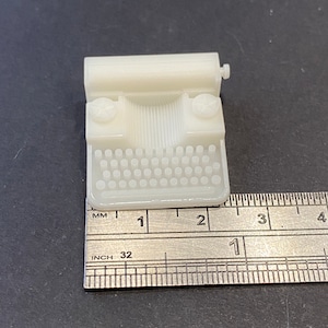May include: A white plastic miniature typewriter measuring approximately 1 inch in length. The typewriter has a small roller, keys, and a carriage return lever.