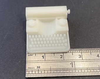 May include: A white plastic miniature typewriter measuring approximately 1 inch in length. The typewriter has a small roller, keys, and a carriage return lever.