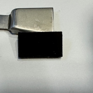 May include: A metal tool with a rectangular black pad. The tool has a silver, rectangular handle and a flat, rectangular head. The black pad is attached to the head. The tool is likely used for a specific craft or repair.