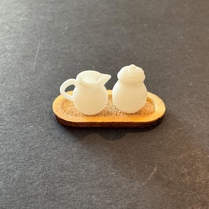 May include: A miniature white creamer and sugar bowl set on a small wooden tray. The creamer has a handle and spout, while the sugar bowl has a lid. The tray is oval-shaped with a textured surface, perfect for a dollhouse or miniature collection.